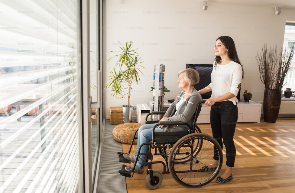 Disabled senior woman in wheelchair at home in her living room, with her young daughter caring for her, looking out the window.